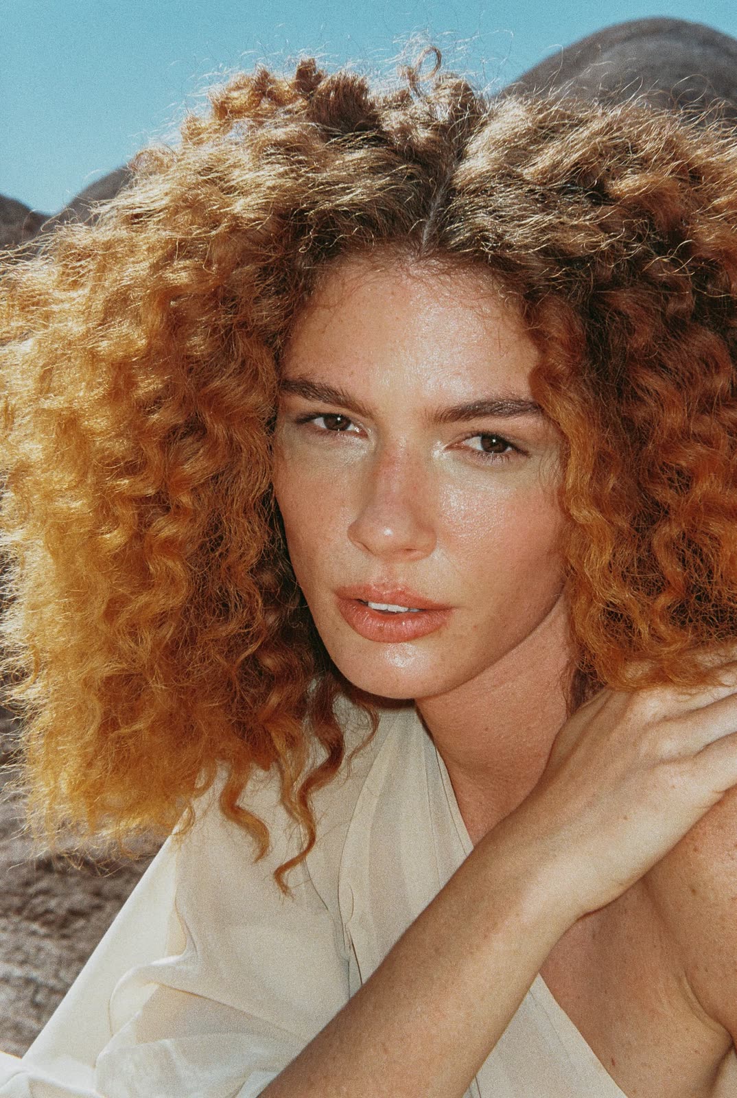 Portrait with red curls in a cream top, desert hills behind — MASCAVO, by Kenneth Kemp