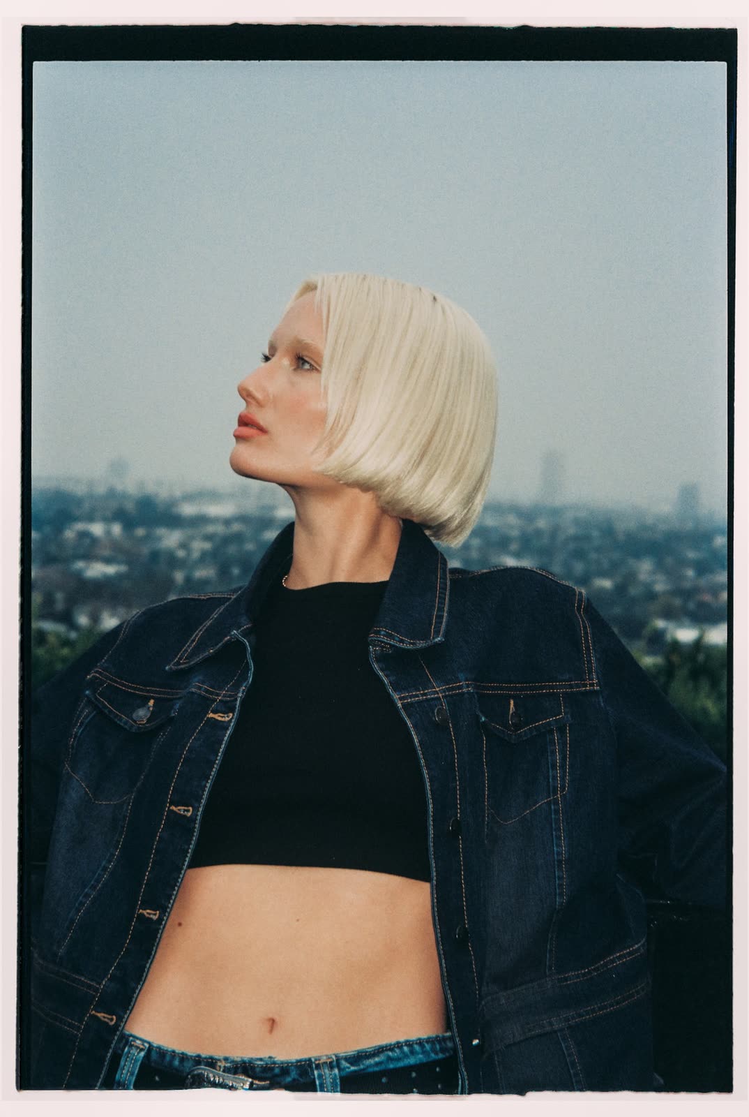 Blonde model in a denim jacket and black crop top, city skyline behind — HAYLEE, by Kenneth Kemp